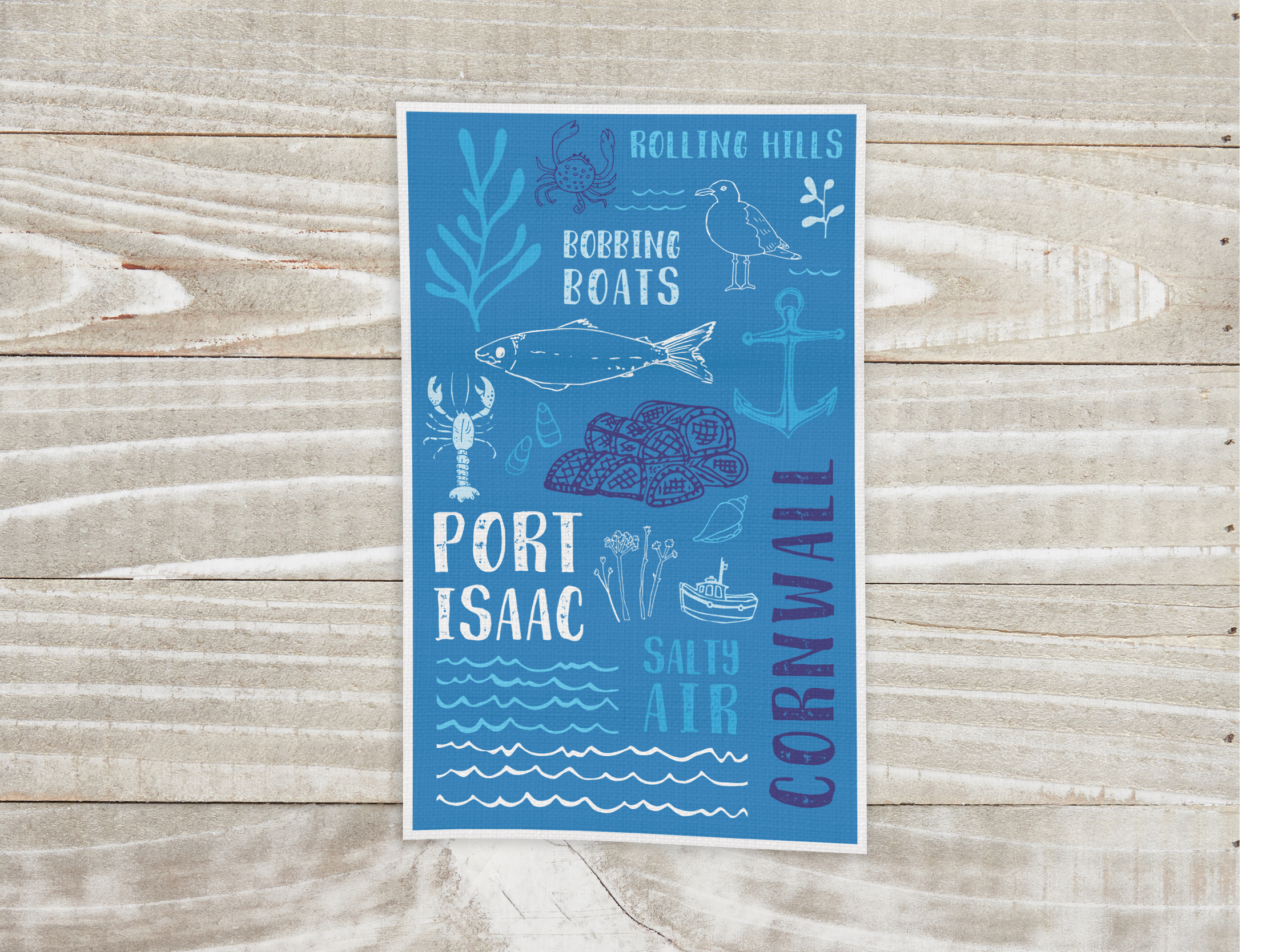 Blue illustrated Port Isaac tea towel displayed on a pale wooden background